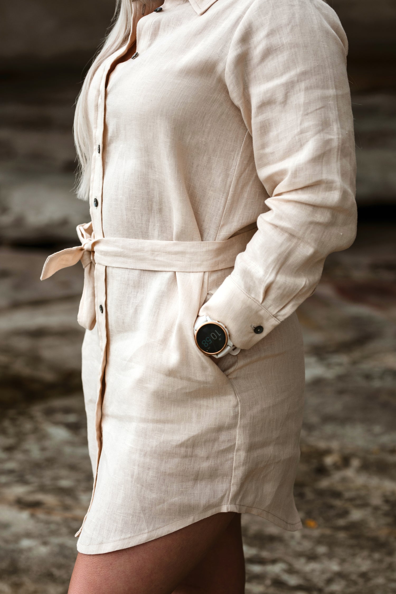 A close up of a petite woman wearing a short, sandy beige shirt dress with a waist tie.