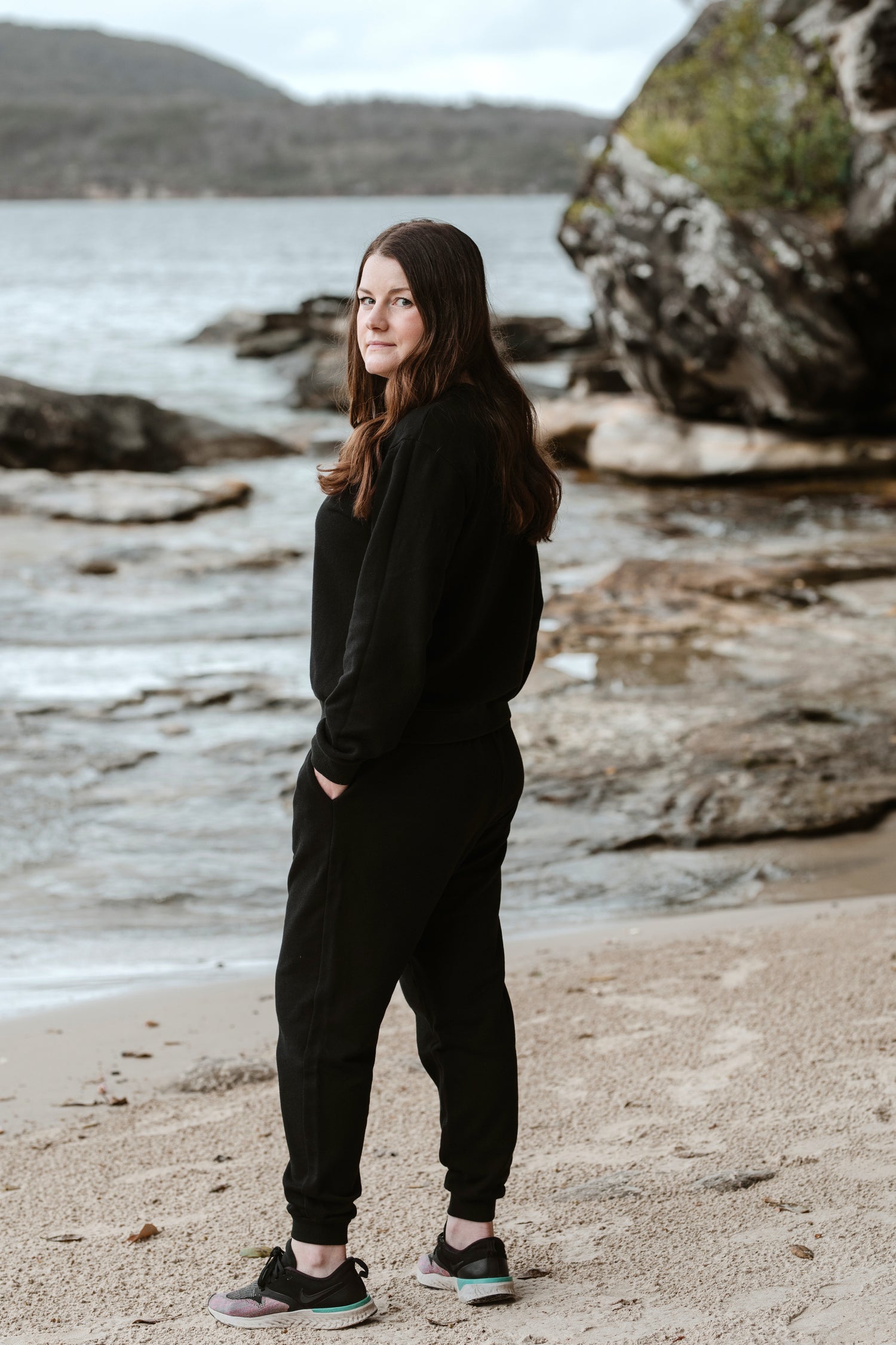 A short woman standing on a beach turning to face the camera. She is wearing a black tracksuit and sneakers.