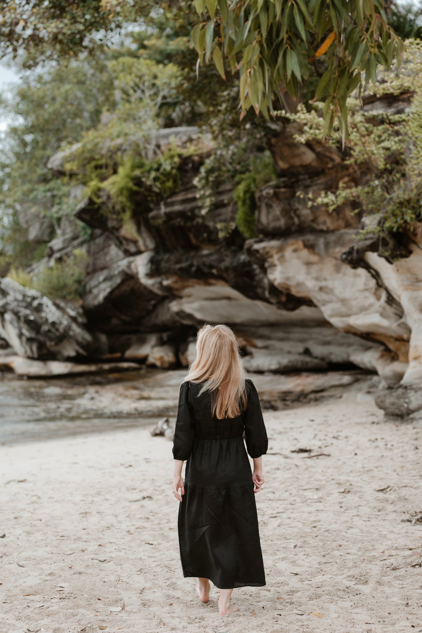 A petite woman walking on a beach with her back to the camera. She is wearing a long black linen dress.