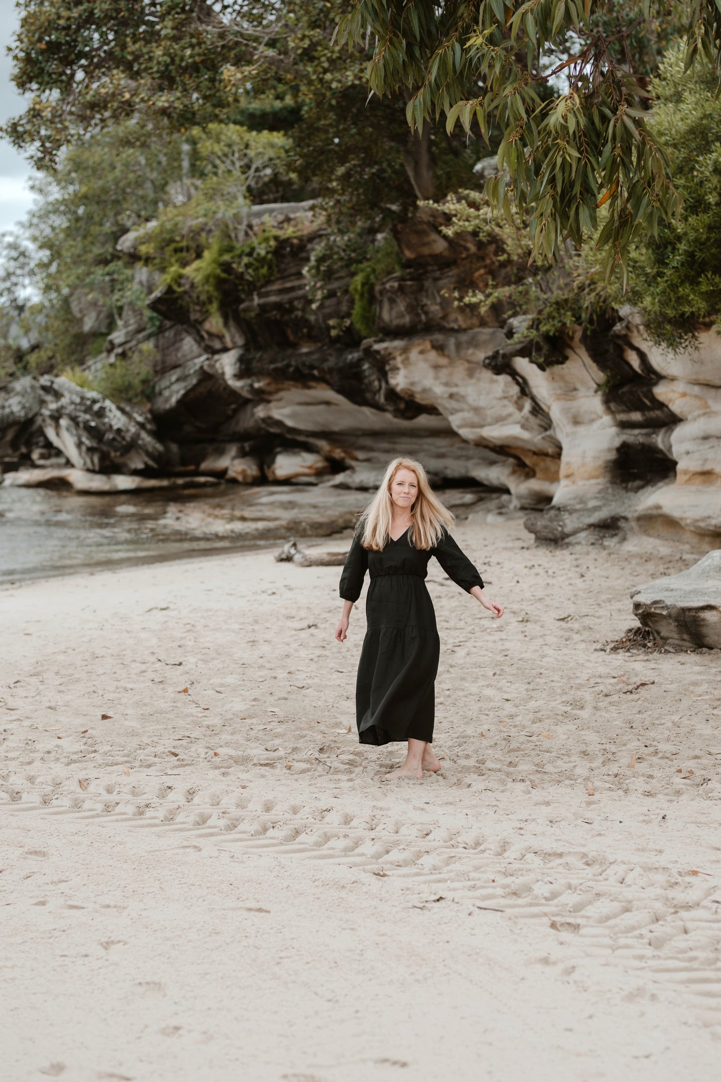 A petite woman on a beach, turning to face the camera. She is wearing a long black linen dress.