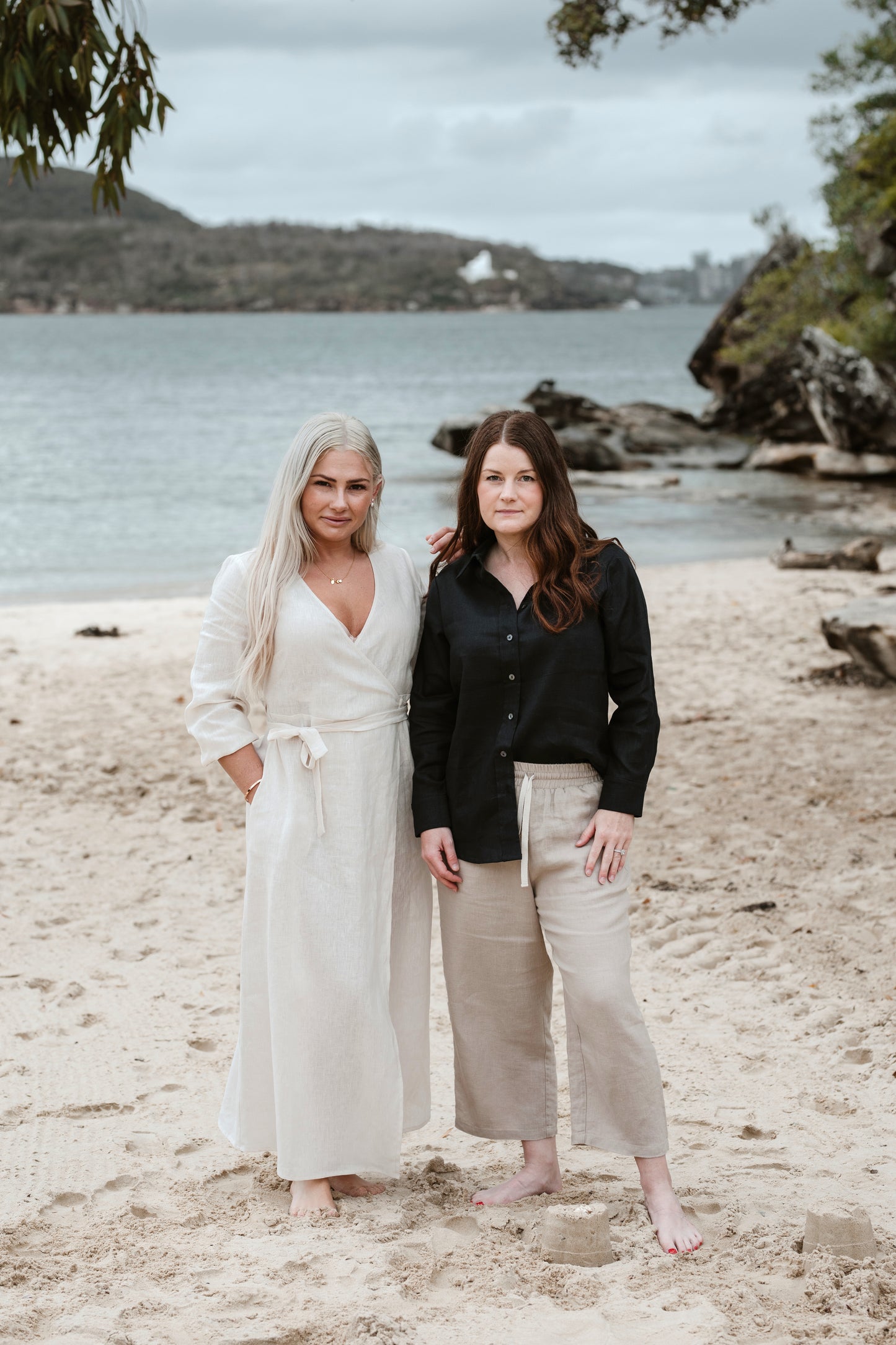 Two petite women on a beach. One wearing an off white linen wrap dress and the other in a navy linen shirt and beige linen pants.